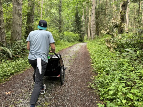 Parent pushing a stroller along a wide hiking trail through forest at Cama Beach State Park