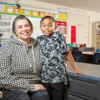 Woman sits in a kid's classroom with an elementary school kid posed next to her.