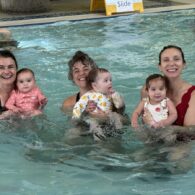 Three women holding babies in front of them, all facing the camera while standing in a pool.