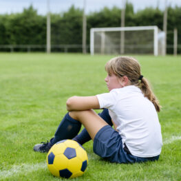 Girl in a soccer uniform sitting on the sidelines with a ball, reflecting after the game.