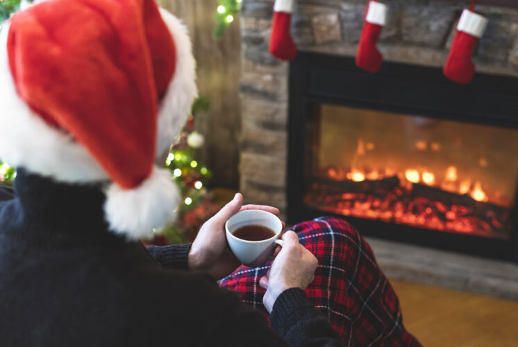 A person wearing a Santa hat sits alone by a glowing fireplace, holding a warm mug, capturing the quiet emotions of spending the holidays in a separated household.
