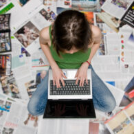 A student sits cross-legged on the floor surrounded by printed newspaper pages, typing on a laptop — symbolizing student journalism and creativity in Washington schools.